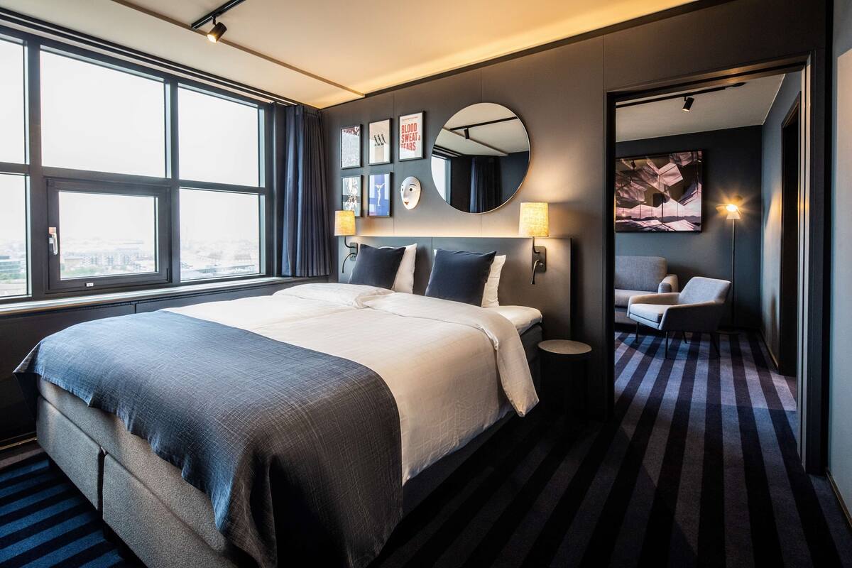 Habitación de hotel moderna con una cama grande, un espejo redondo y vistas a la ciudad a través de amplias ventanas.