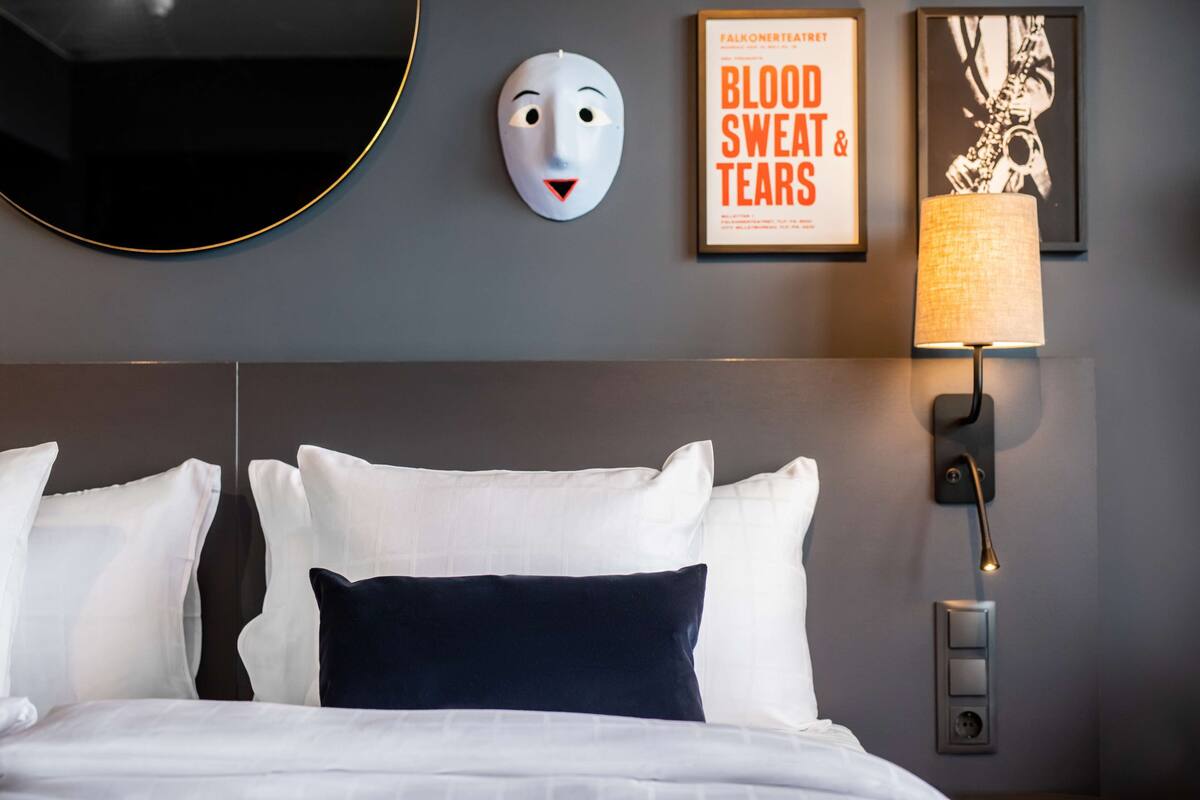 Una habitación de hotel con una cama bien hecha, una lámpara de pared, un cuadro enmarcado y una máscara decorativa.