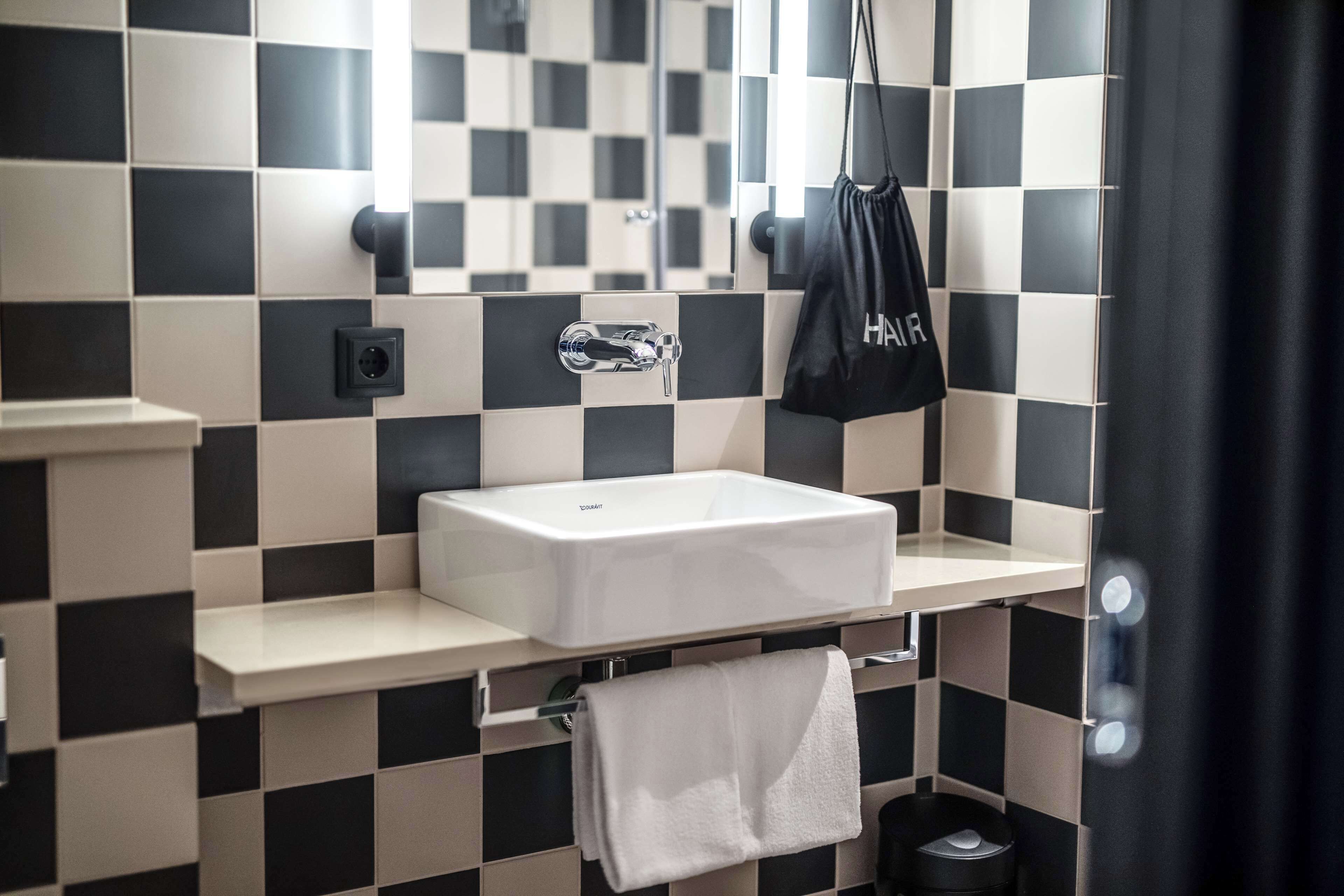 Un baño moderno con un patrón de baldosas blanco y negro a cuadros, un lavabo blanco, una toalla colgada en un perchero y un bolso negro con la palabra 'HAIR'.