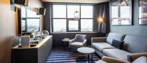 Habitación de hotel moderna con un ventanal grande, un sofá, una mesa de centro, una silla y un escritorio con cafetera.