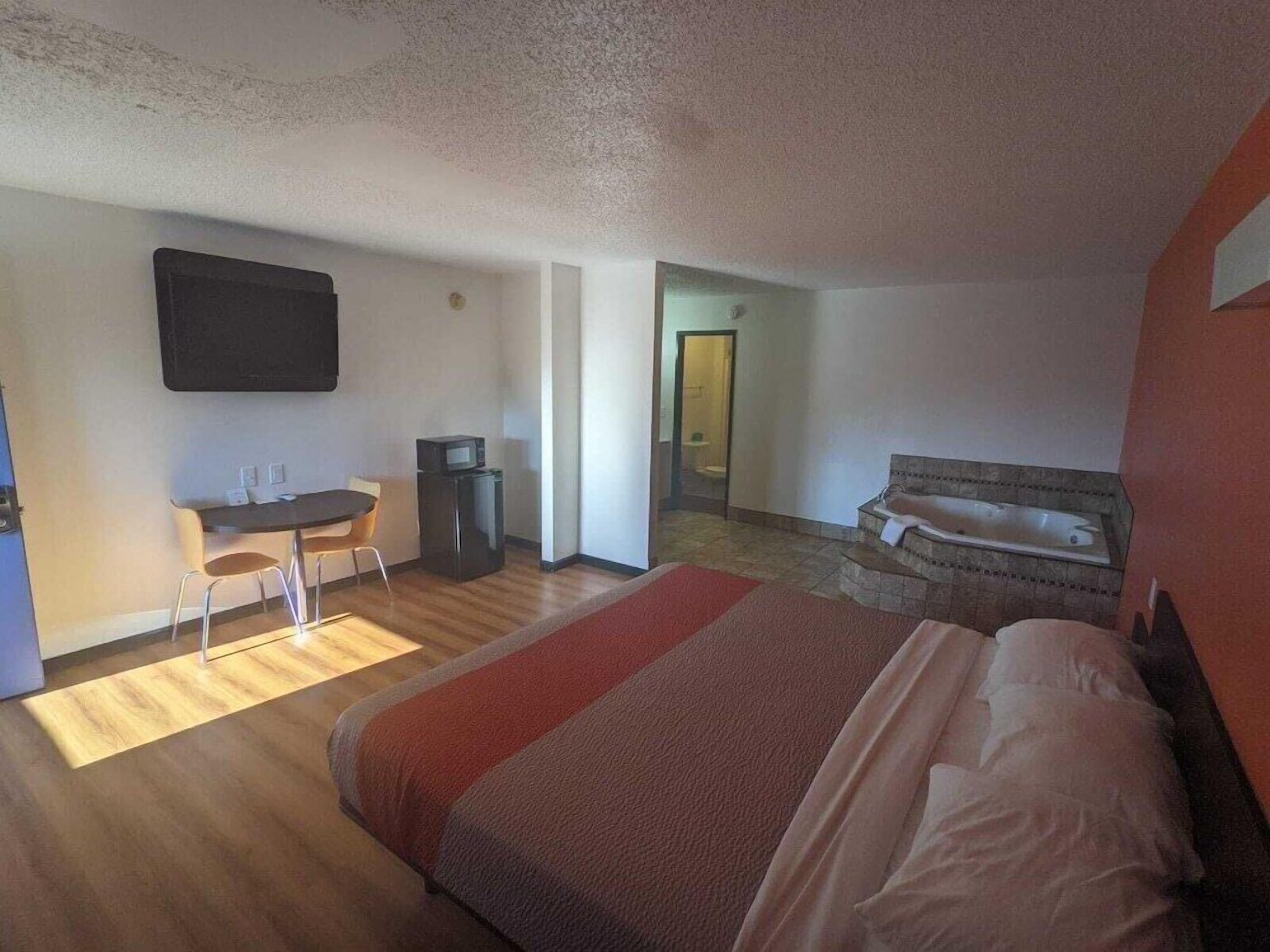 A hotel room with a bed, a desk with a chair, a television, and a bathroom visible through an open door.