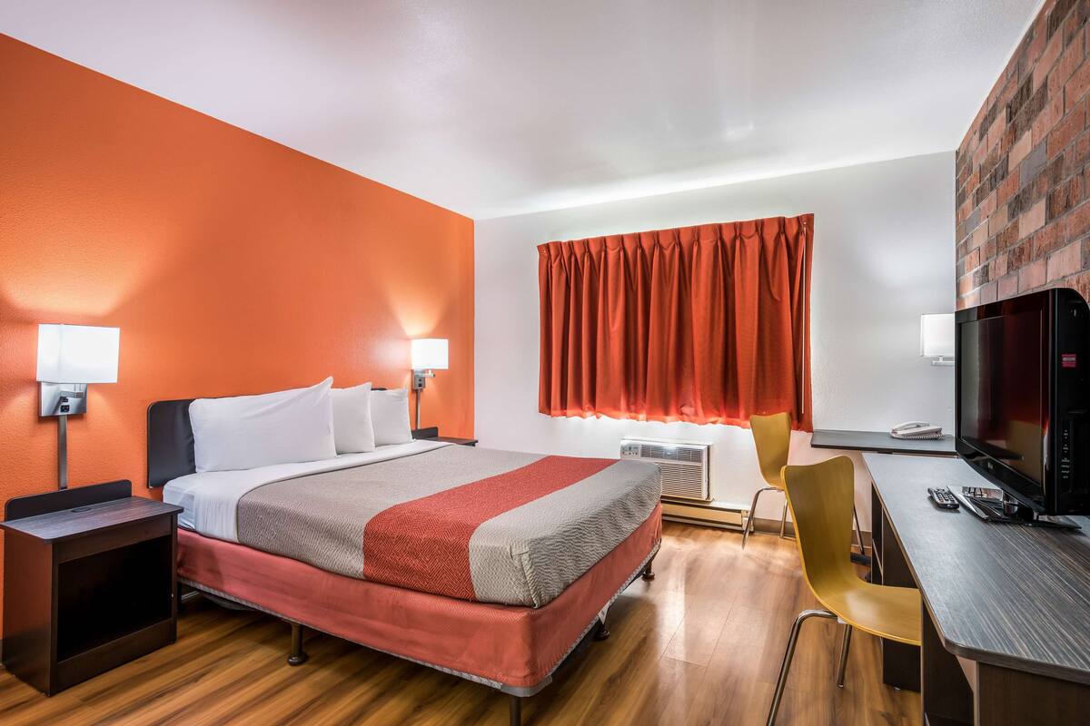 A hotel room with a bed, bedside tables, a desk, a chair, a television, and a window with curtains.