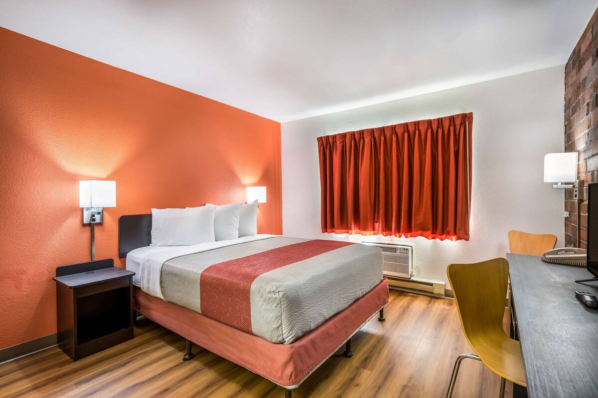 A hotel room with a bed, bedside tables, a desk with a chair, a window with curtains, and a brick wall.
