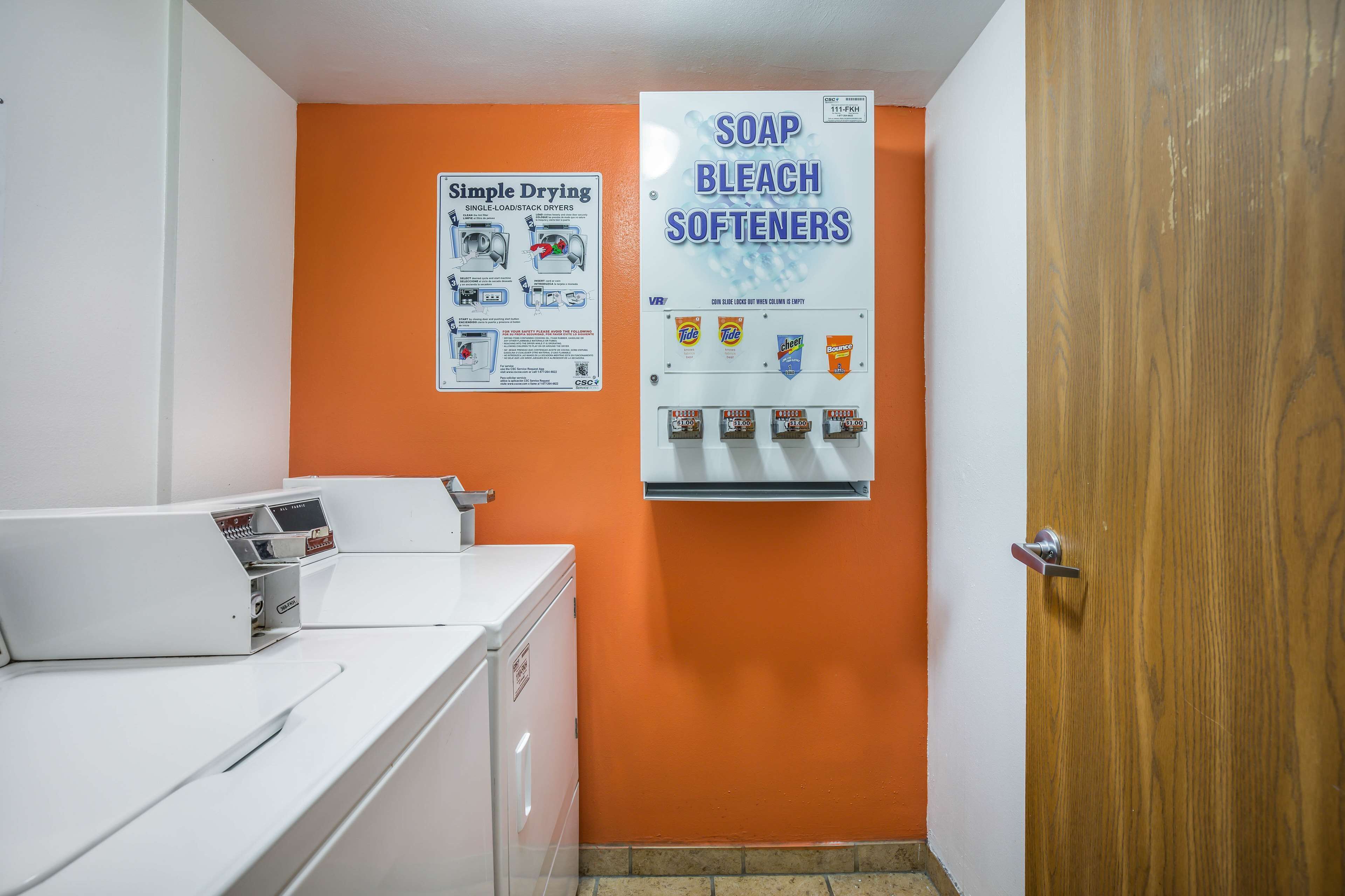 A laundry area with a white washing machine, a wall-mounted dryer, and a bulletin board advertising laundry products.