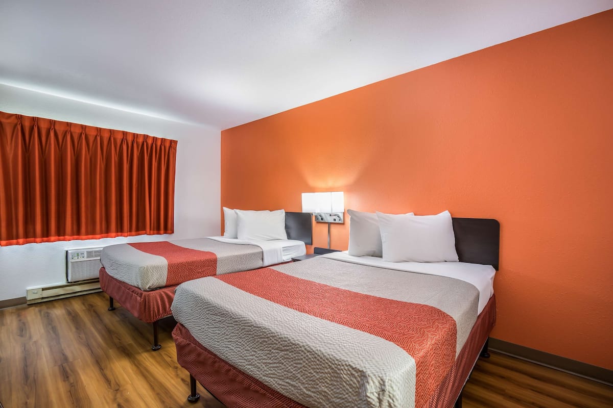 A hotel room with two beds, orange walls, wooden flooring, and a window with red curtains.