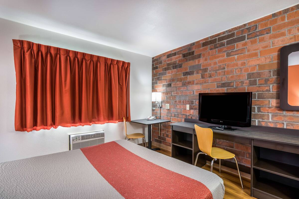 A hotel room with a bed, a desk, a TV, and a brick wall.