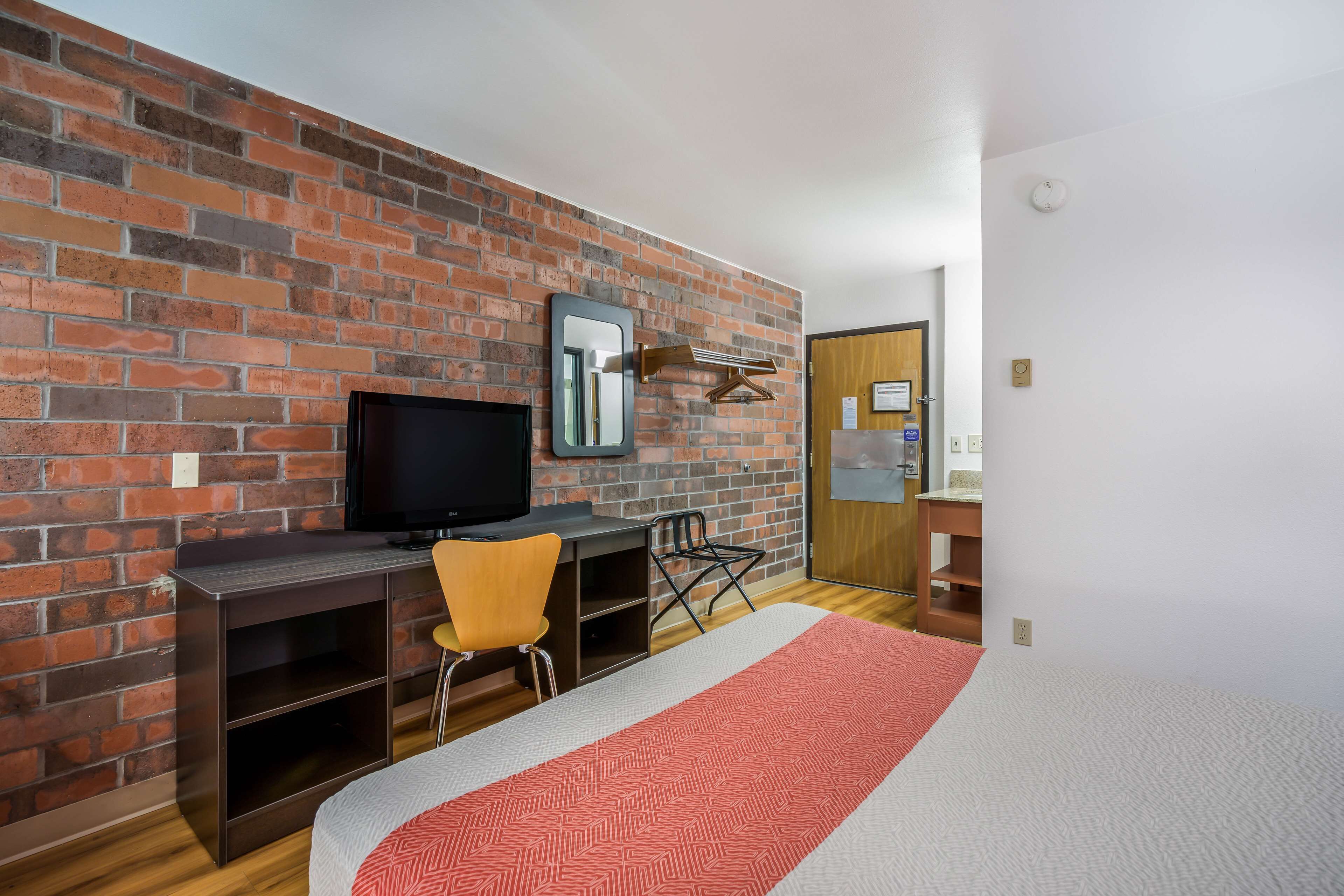 Ein Hotelzimmer mit Backsteinwand, einem Flachbildfernseher, einem Schreibtisch mit Stuhl, einem Bett mit gemustertem Überwurf und einer Holztür.