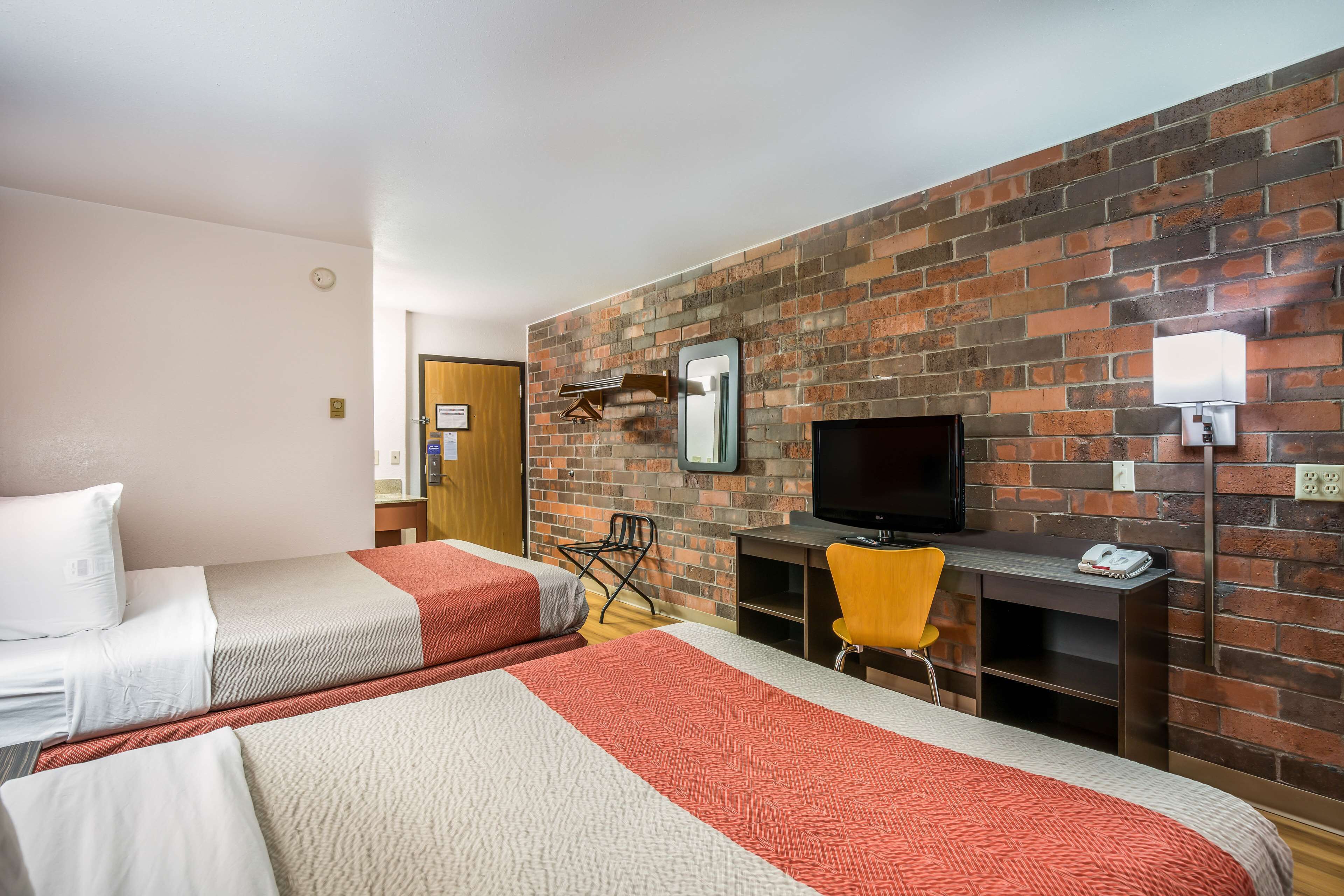 A hotel room with two beds, a TV, a desk, and a brick wall.