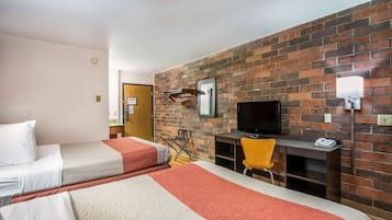 A hotel room with two beds, a TV, a desk, and a brick wall.