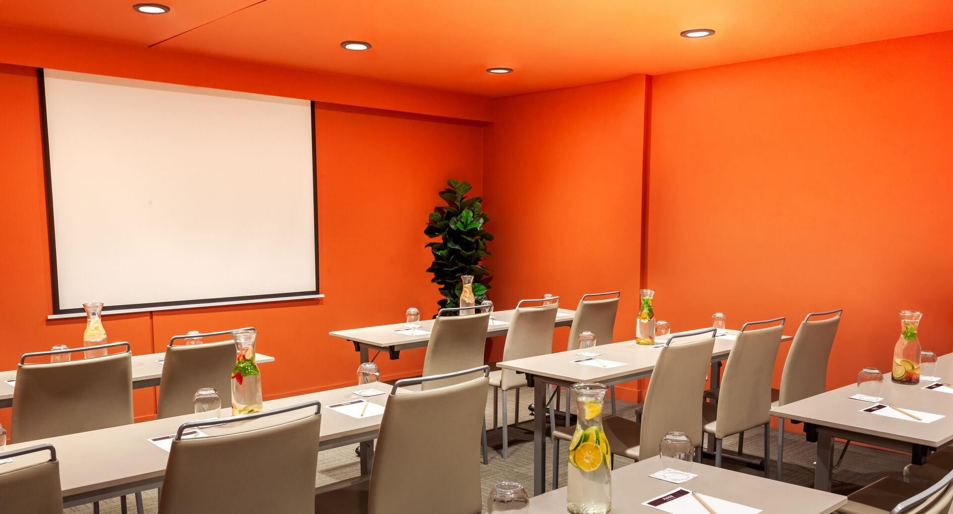 Un salón de conferencias con mesas, sillas, una pantalla de proyector y una planta en maceta.