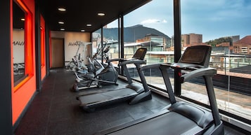 Un gimnasio con cintas de correr y equipo de ejercicio, ventanas amplias con vista a la ciudad y una pared de acento rojo.