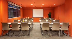 Un salón de conferencias con mesas y sillas, una pantalla de proyector y un ventanal con persianas.