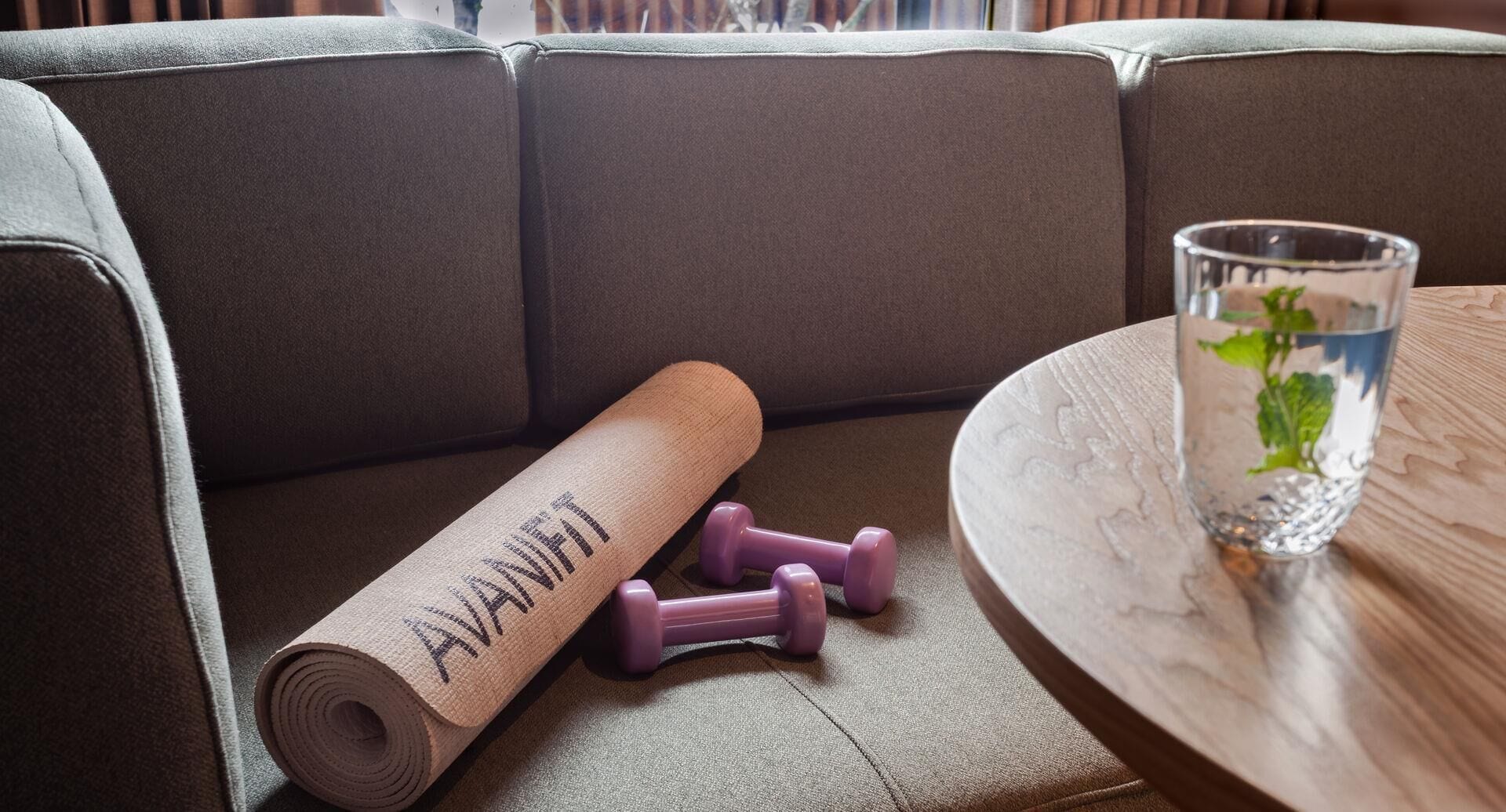 Eine Yogamatte mit dem Aufdruck 'AVANTIK', zwei violette Kurzhanteln und ein Glas Wasser mit einem Minzipflanzchen auf einem Holztisch.