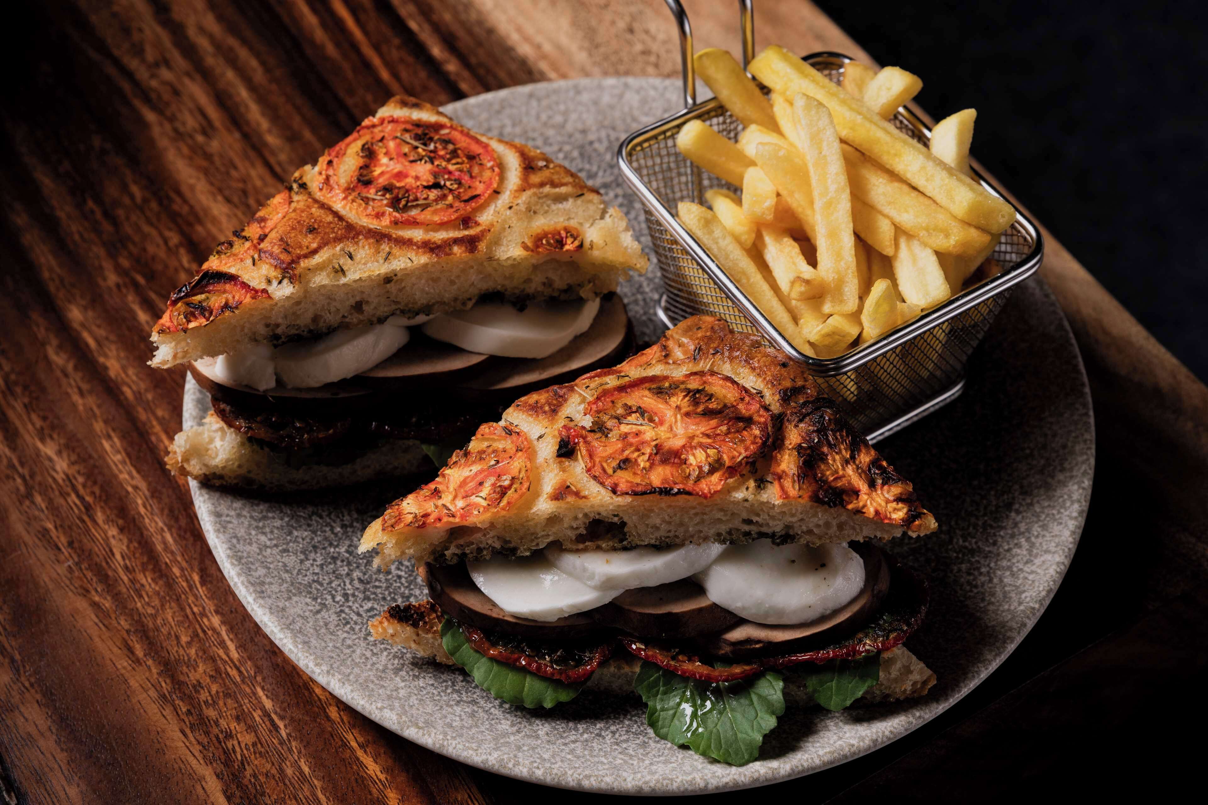 Un sandwich garni de tranches de tomates grillées, servi sur une assiette, accompagné d’un panier de frites.