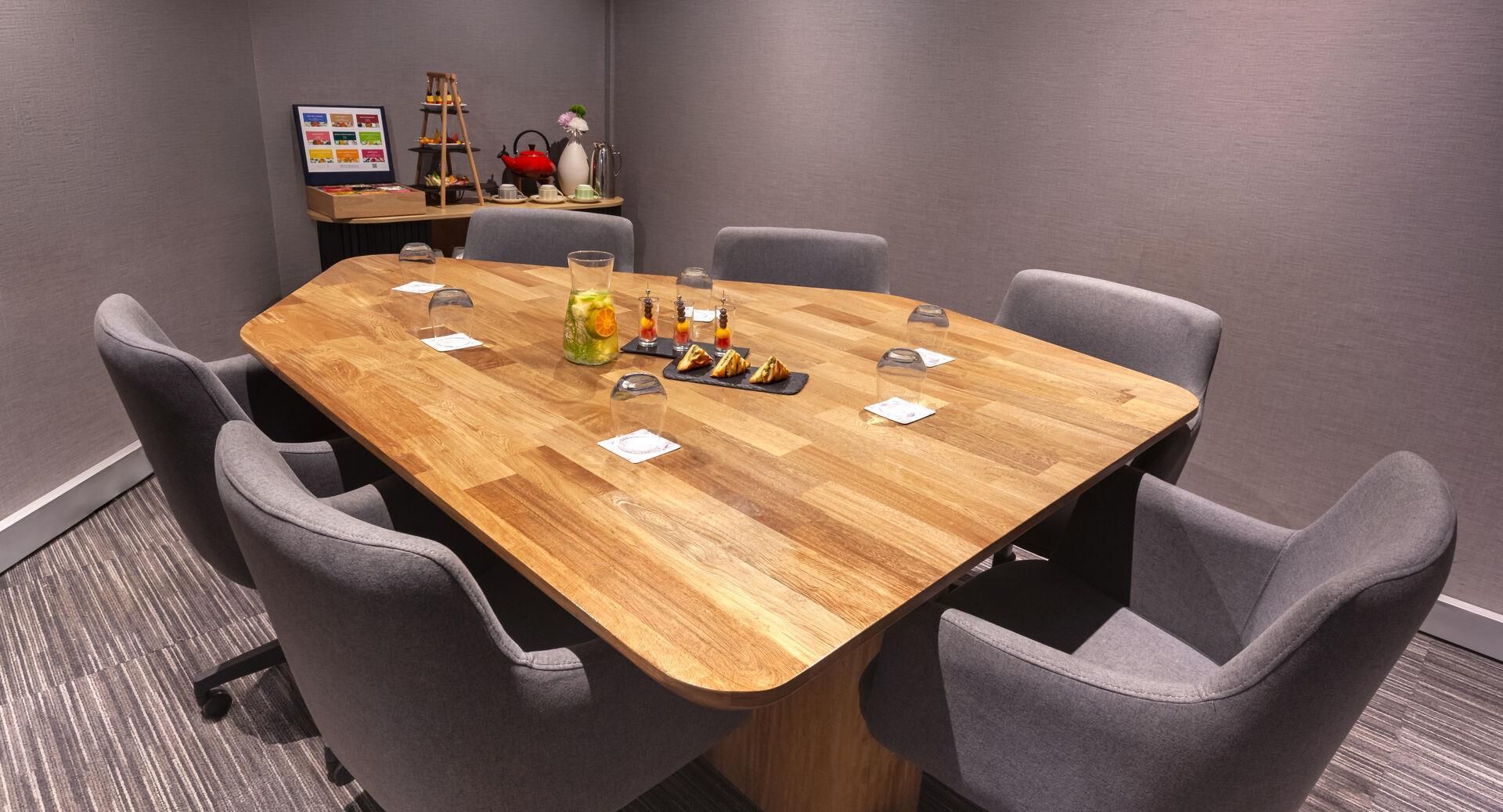 A conference room with a wooden table, grey upholstered chairs, and a small display of snacks and drinks.