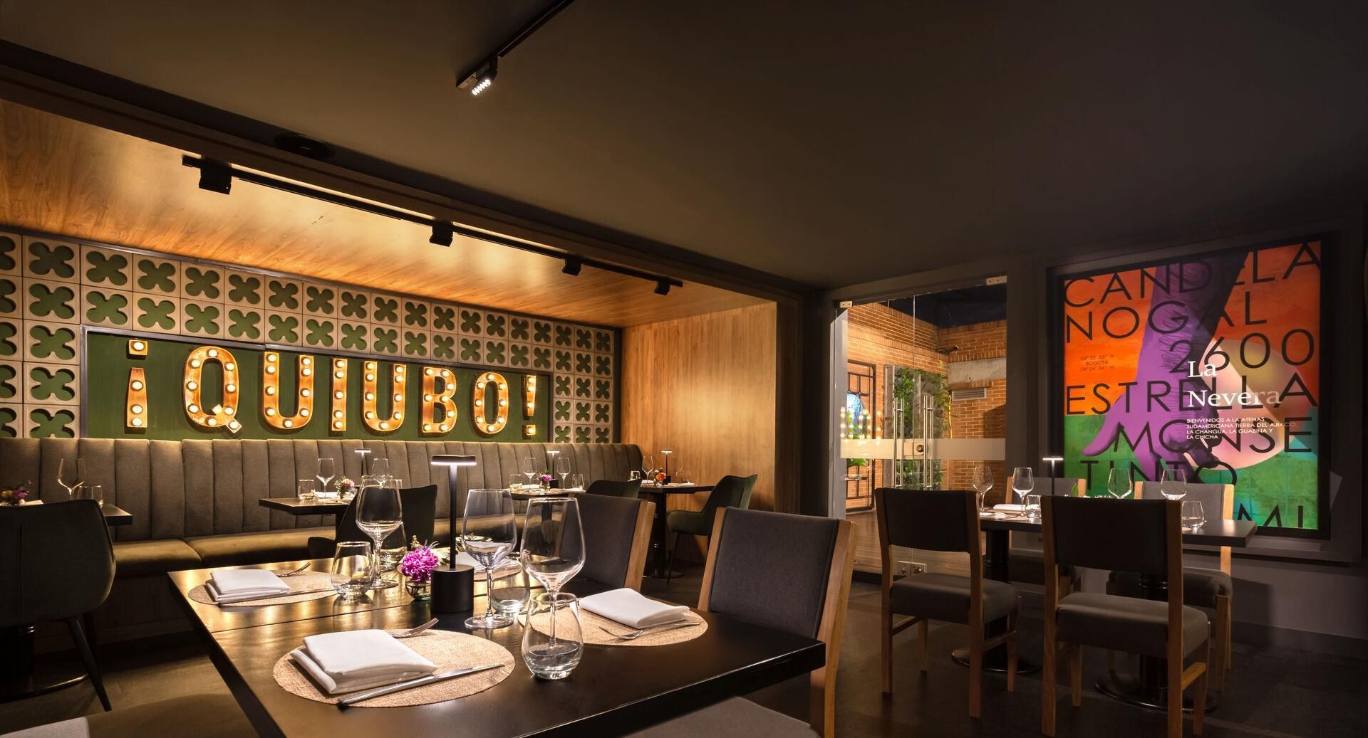 A restaurant interior with a prominent illuminated sign reading 'QUIUBO!' and a colorful mural on the wall.