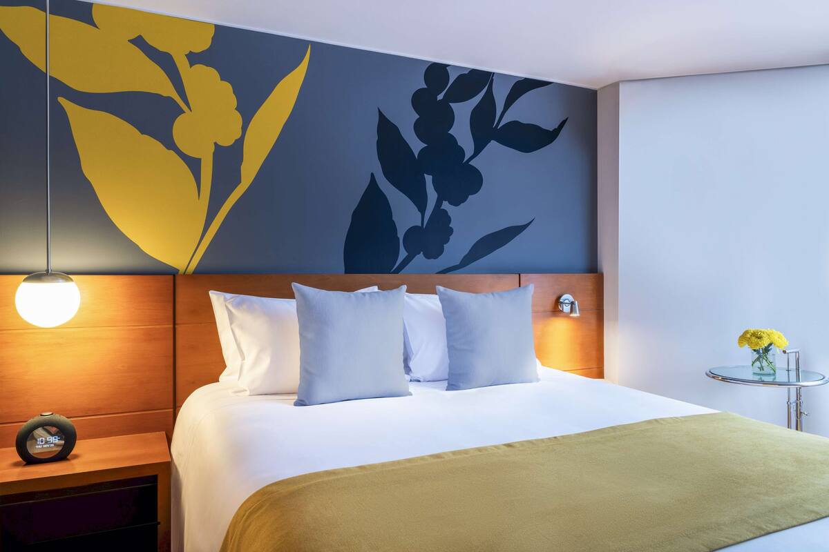 Una habitación de hotel con una cama grande, una mesita de noche con un reloj, una pared con arte floral y una pequeña mesa con un jarrón de flores.