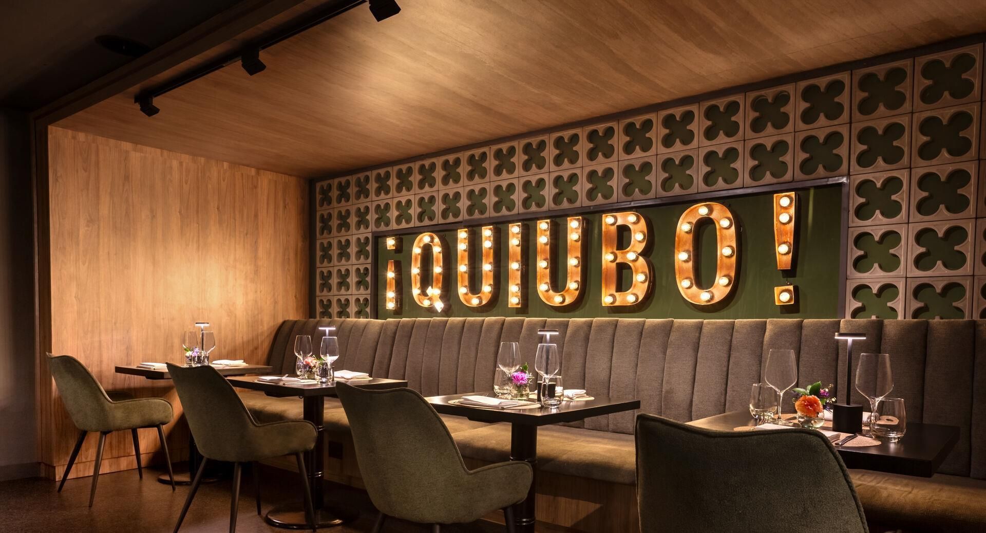 A restaurant interior with a booth seating area, tables with chairs, and a wall with illuminated letters spelling 'QUIUBO!'
