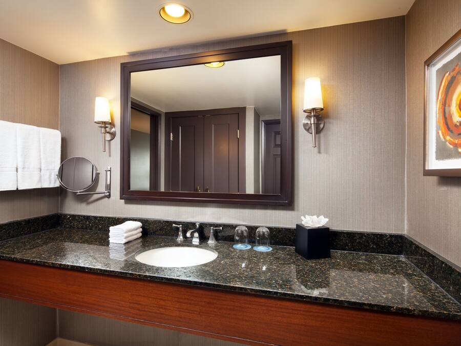 A hotel bathroom with a granite countertop, a round mirror, a white sink, and two wall-mounted lights.
