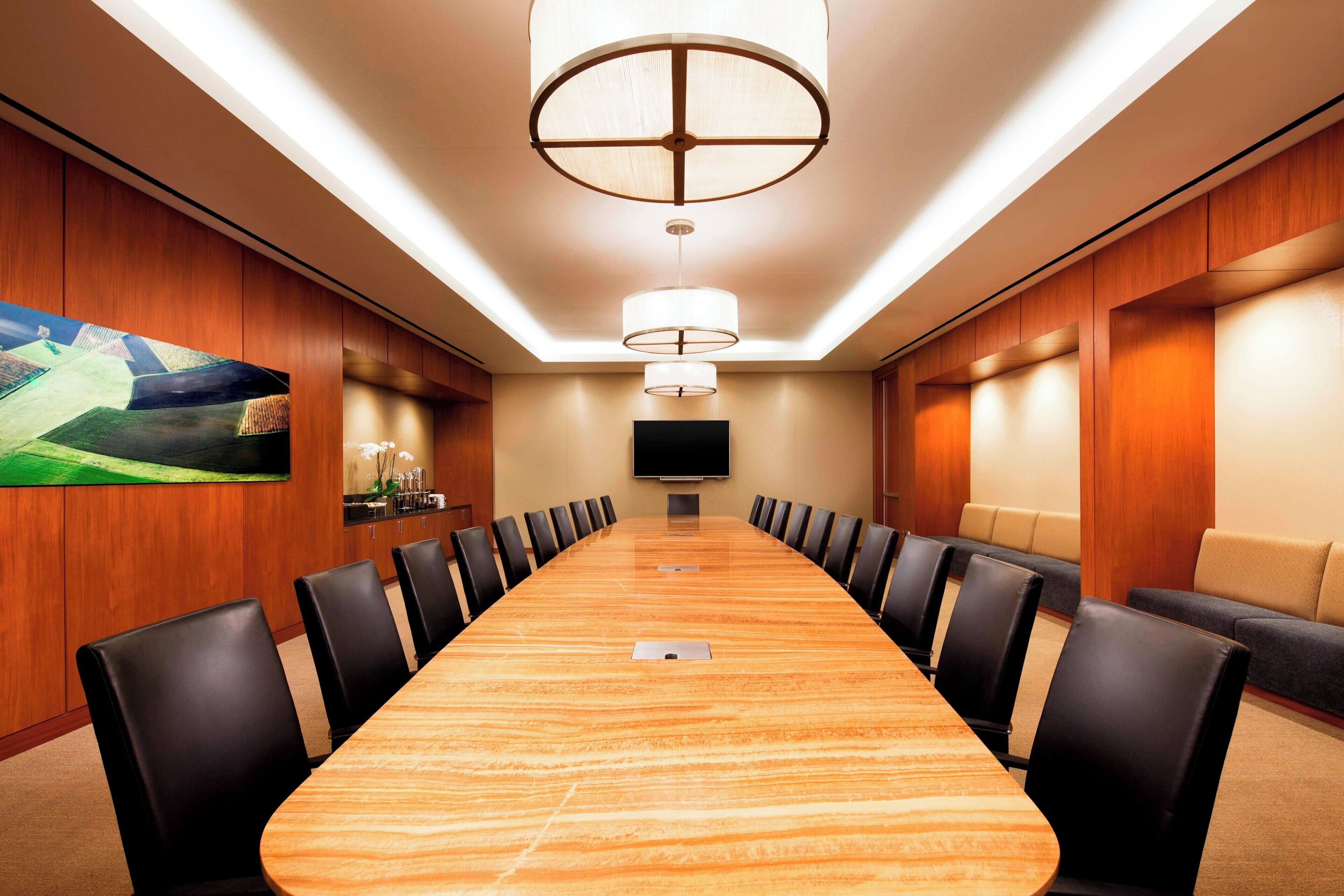 Un salón de conferencias con una larga mesa, sillas negras, un cuadro grande, un televisor de pantalla plana y un accesorio de iluminación en el techo.