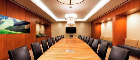 Un salón de conferencias con una larga mesa, sillas negras, un cuadro grande, un televisor de pantalla plana y un accesorio de iluminación en el techo.