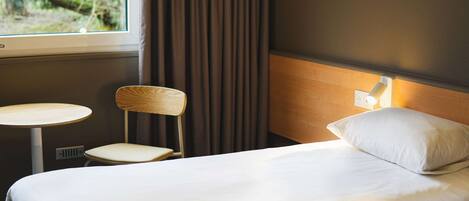 A hotel room with a bed, a chair, a small table, and a window with curtains.