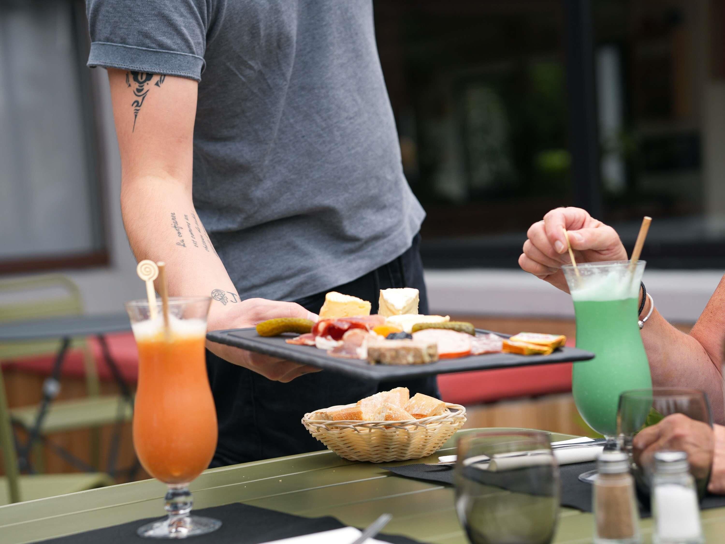 Una persona sirviendo comida y bebidas en una mesa, con un cesto de pan, un vaso de zumo de naranja y una bebida verde.