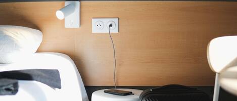 Una habitación de hotel con una cama, una mesita de noche con teléfono, un enchufe y una maleta.