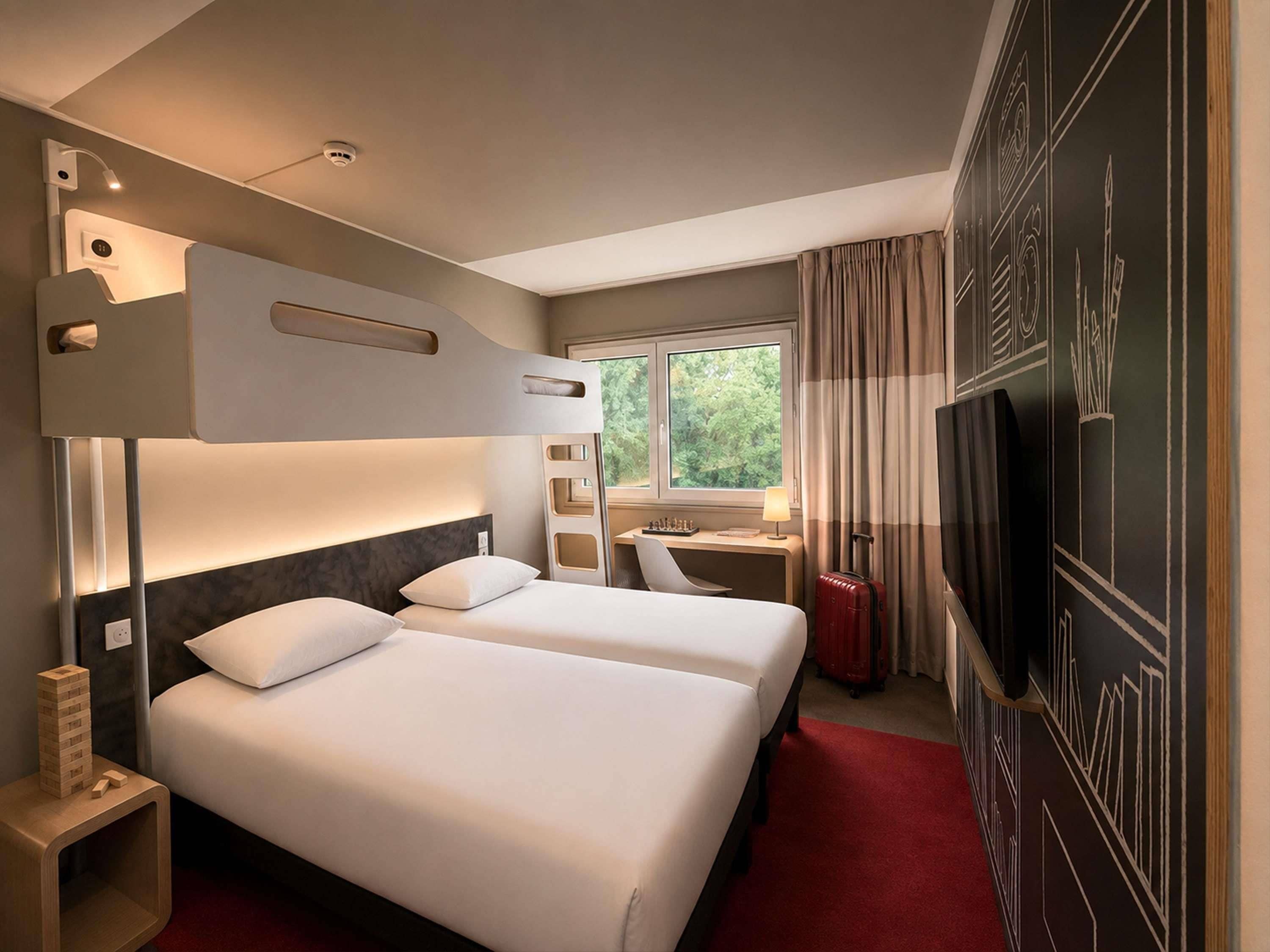 A hotel room with a bed, desk, chair, TV, and a view of greenery outside.