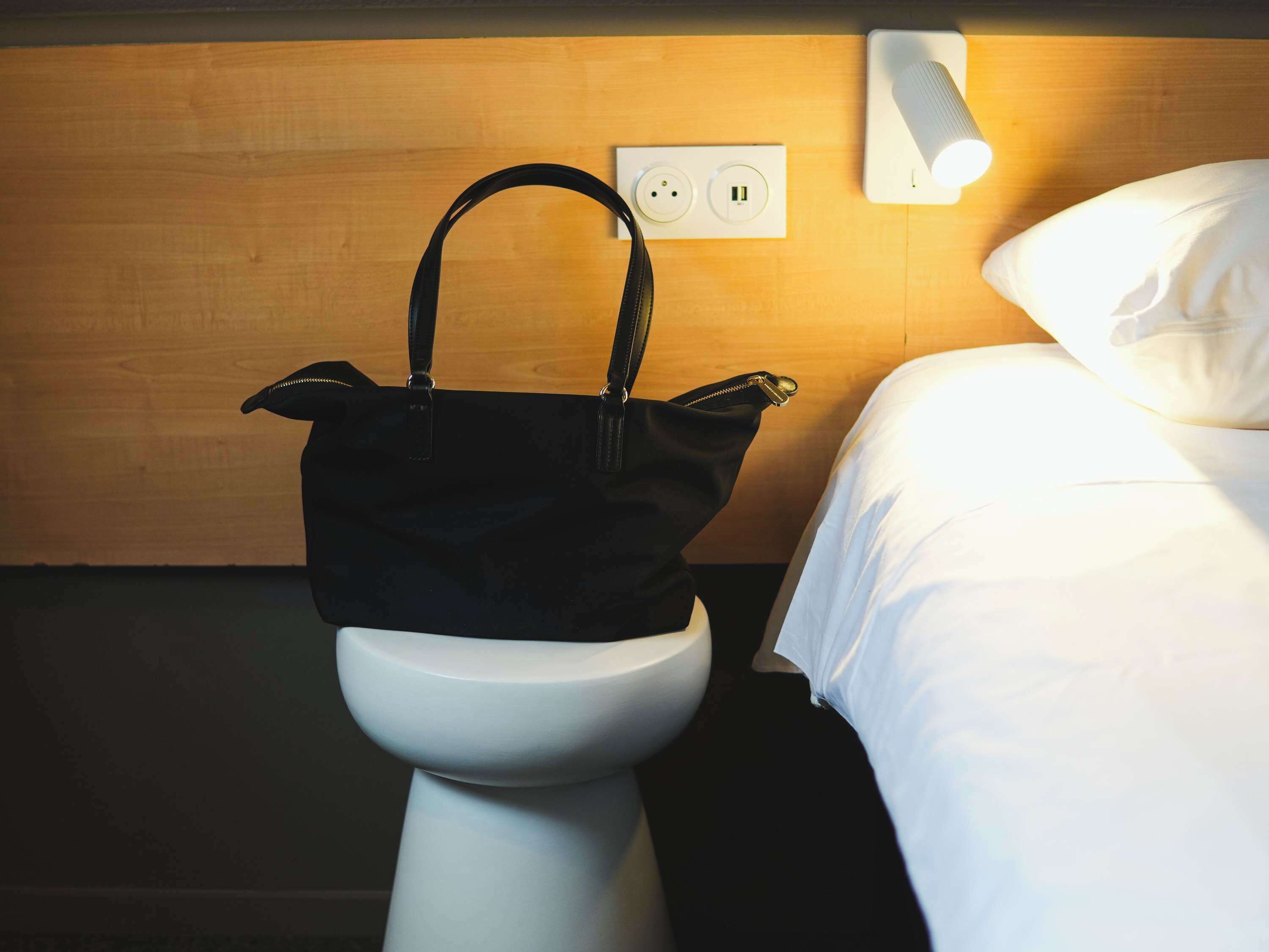 A hotel room with a white toilet, a bedside table, a lamp, and a power outlet on the wall.
