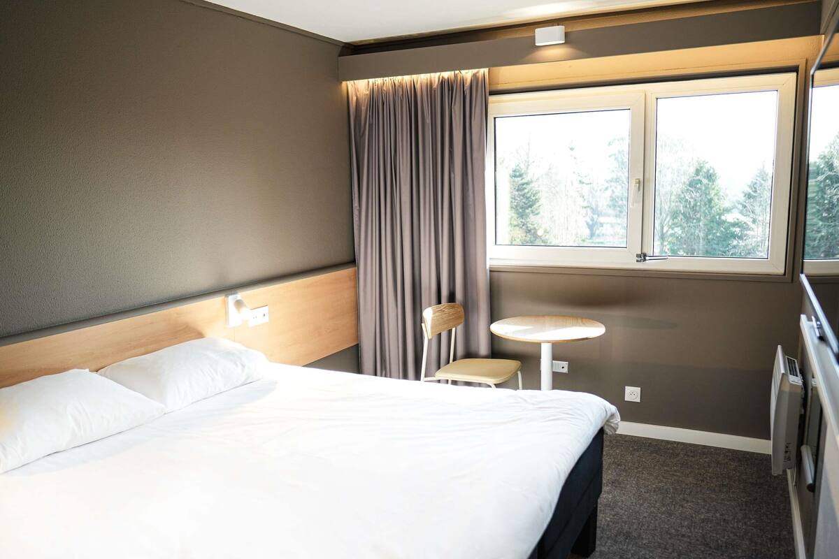 A hotel room with a bed, a small round table, a chair, a window with curtains, and a radiator.