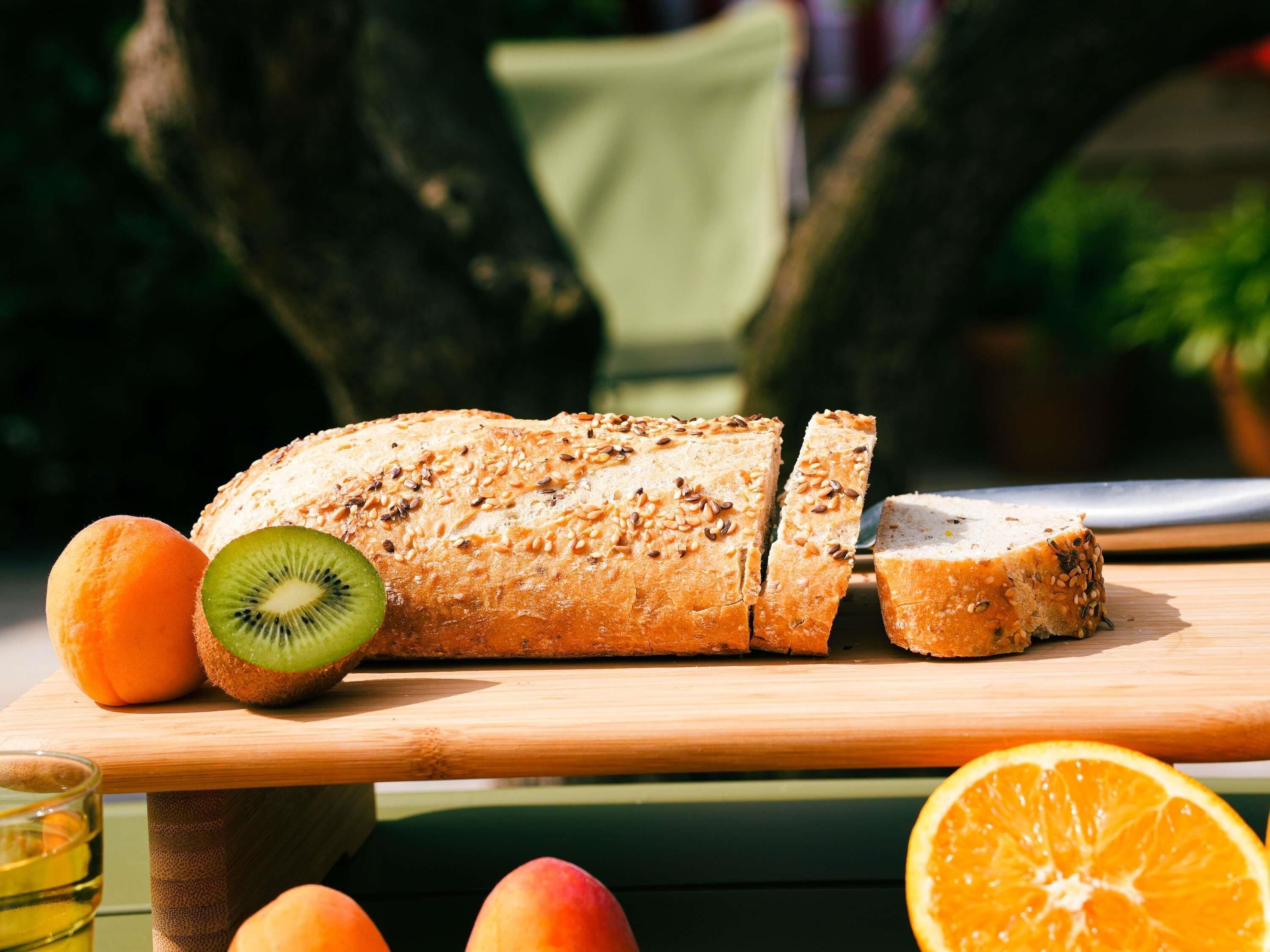 Una hogaza de pan con semillas de sésamo sobre una tabla de madera, acompañada de frutas como kiwi, naranja y melocotón.