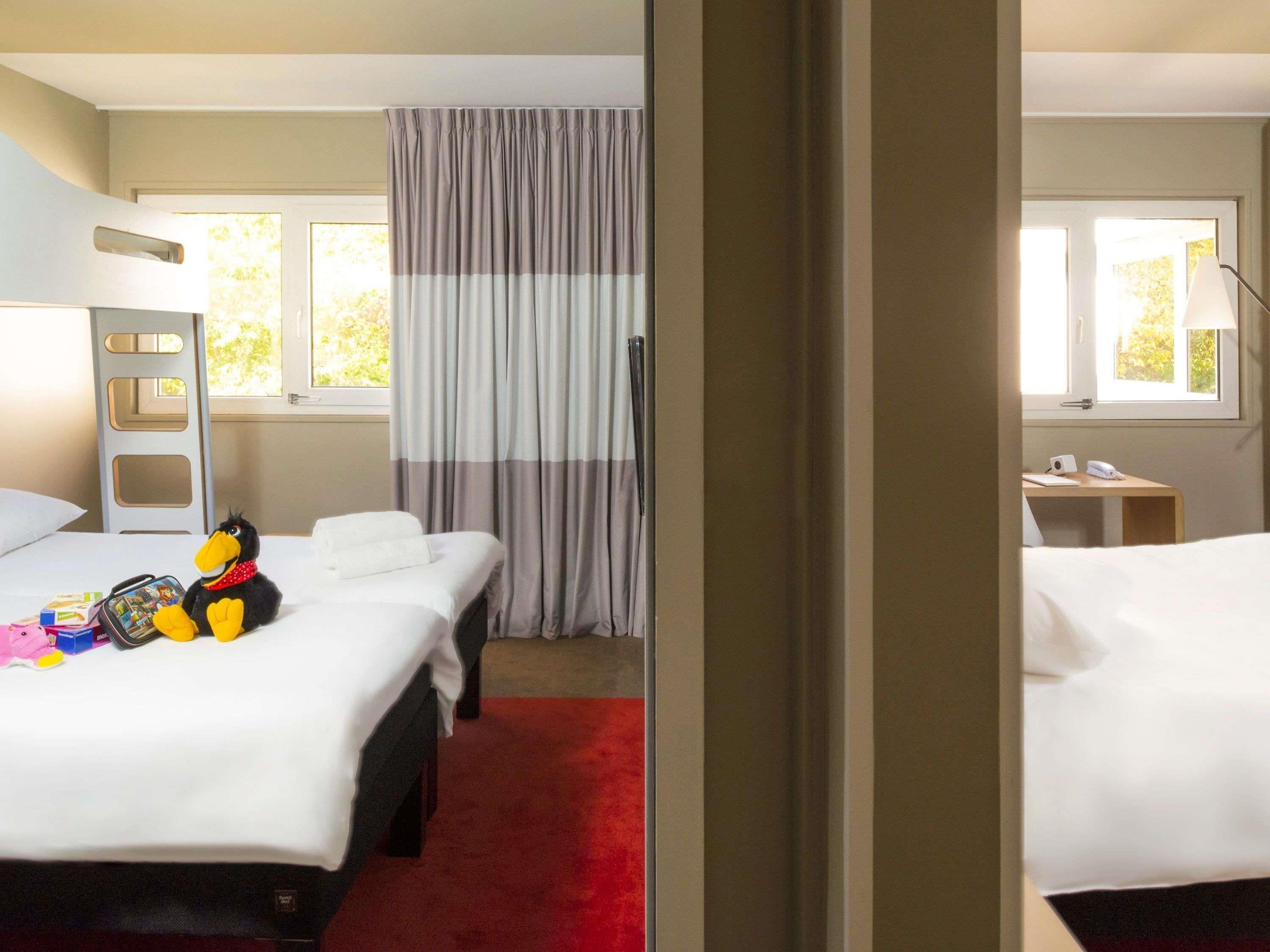 A hotel room with two beds, a red carpet, a window with curtains, and a desk with a lamp.