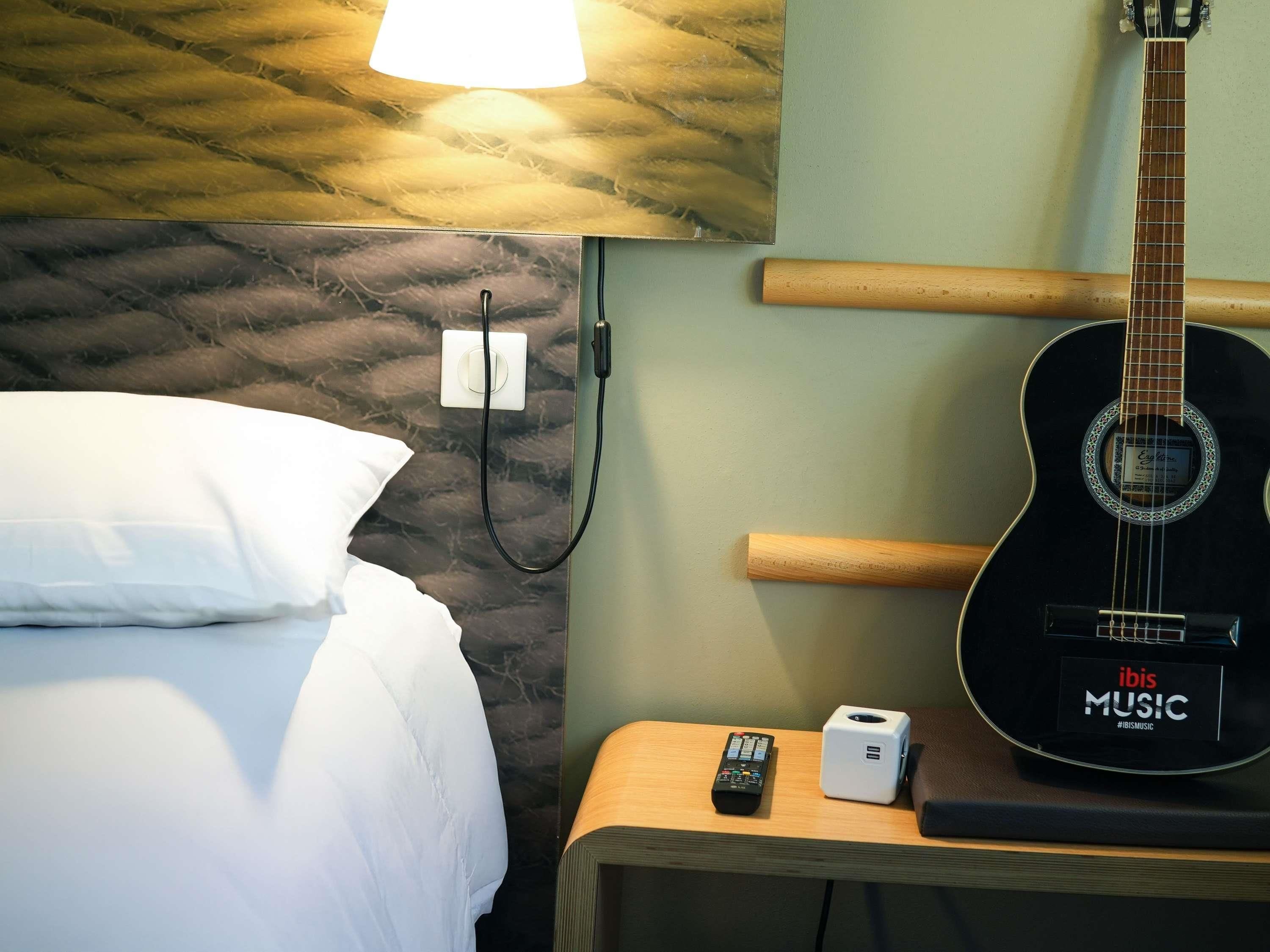 A hotel room with a bed, a guitar, a lamp, a power outlet, and a small table with a remote control and a device.