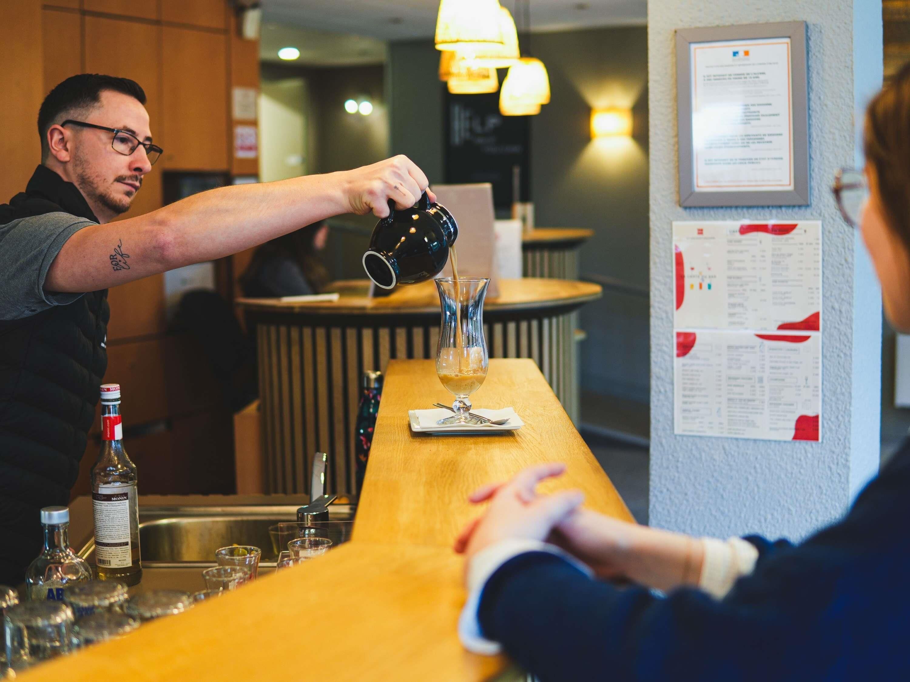 A bar area with a bartender pouring a drink into a glass, a menu on the wall, and a bottle of liquor on the counter.