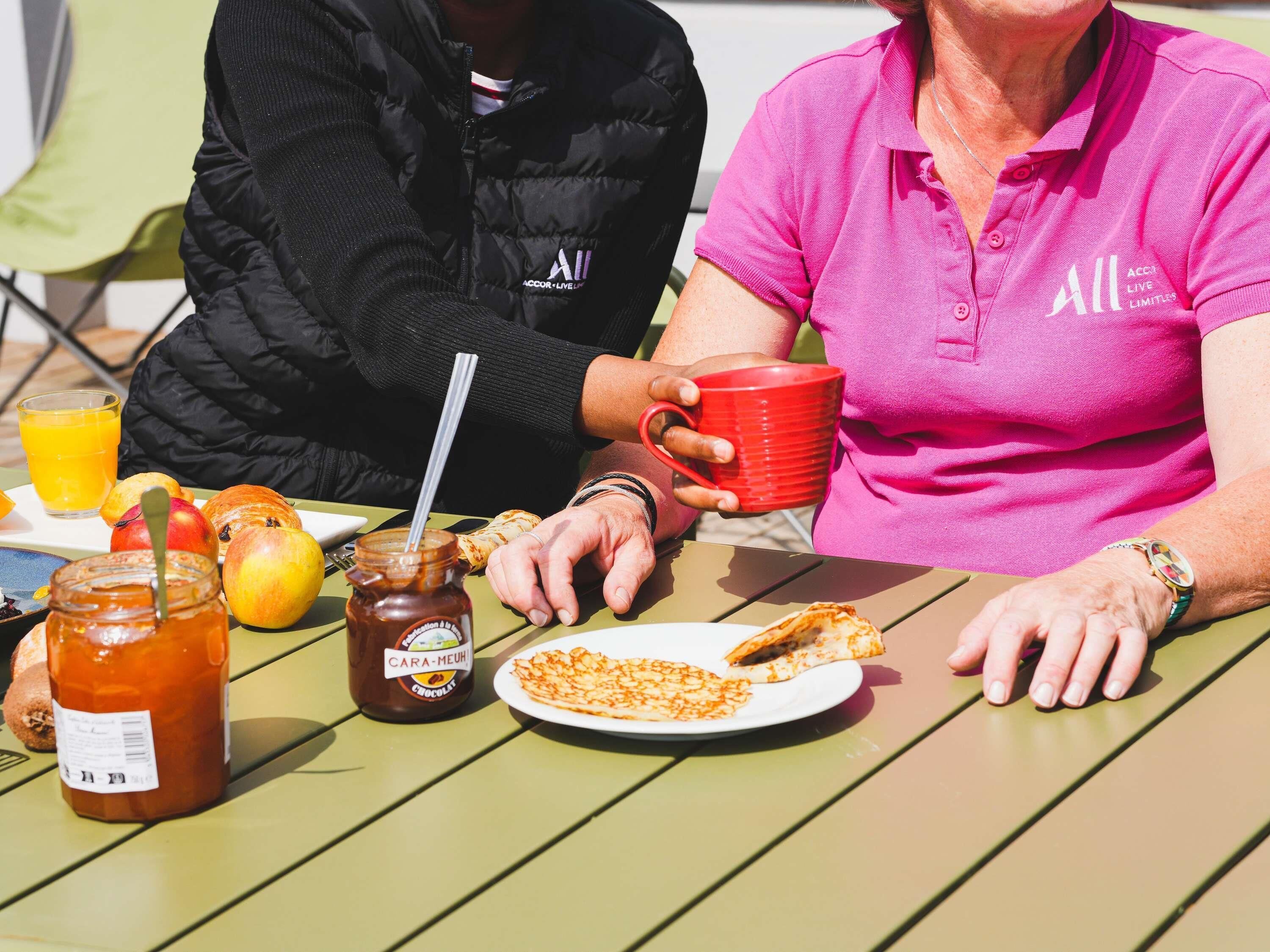 A table with breakfast items including a plate of food, a red mug, a jar of jam, and a glass of orange juice.