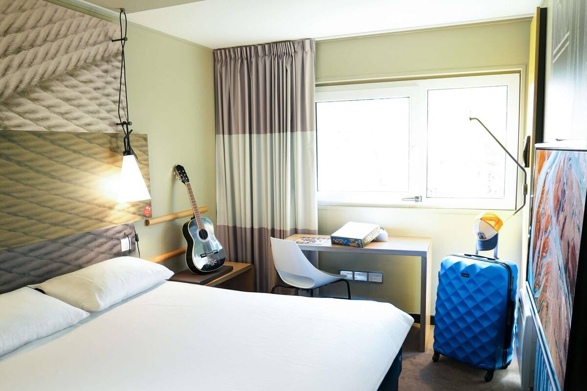 Une chambre d’hôtel avec un lit, un bureau, une chaise, une guitare, une valise et une fenêtre avec des rideaux.