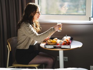 Une personne assise à une table ronde, devant un verre de jus d’orange, un panier de viennoiseries et une tasse de café.