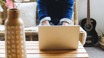 Une personne utilise un ordinateur portable sur une table en bois, avec une chaise en osier et une guitare en arrière-plan.