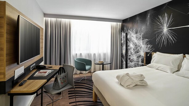 A hotel room with a bed, a desk with a computer, a chair, a TV, and a window with curtains.