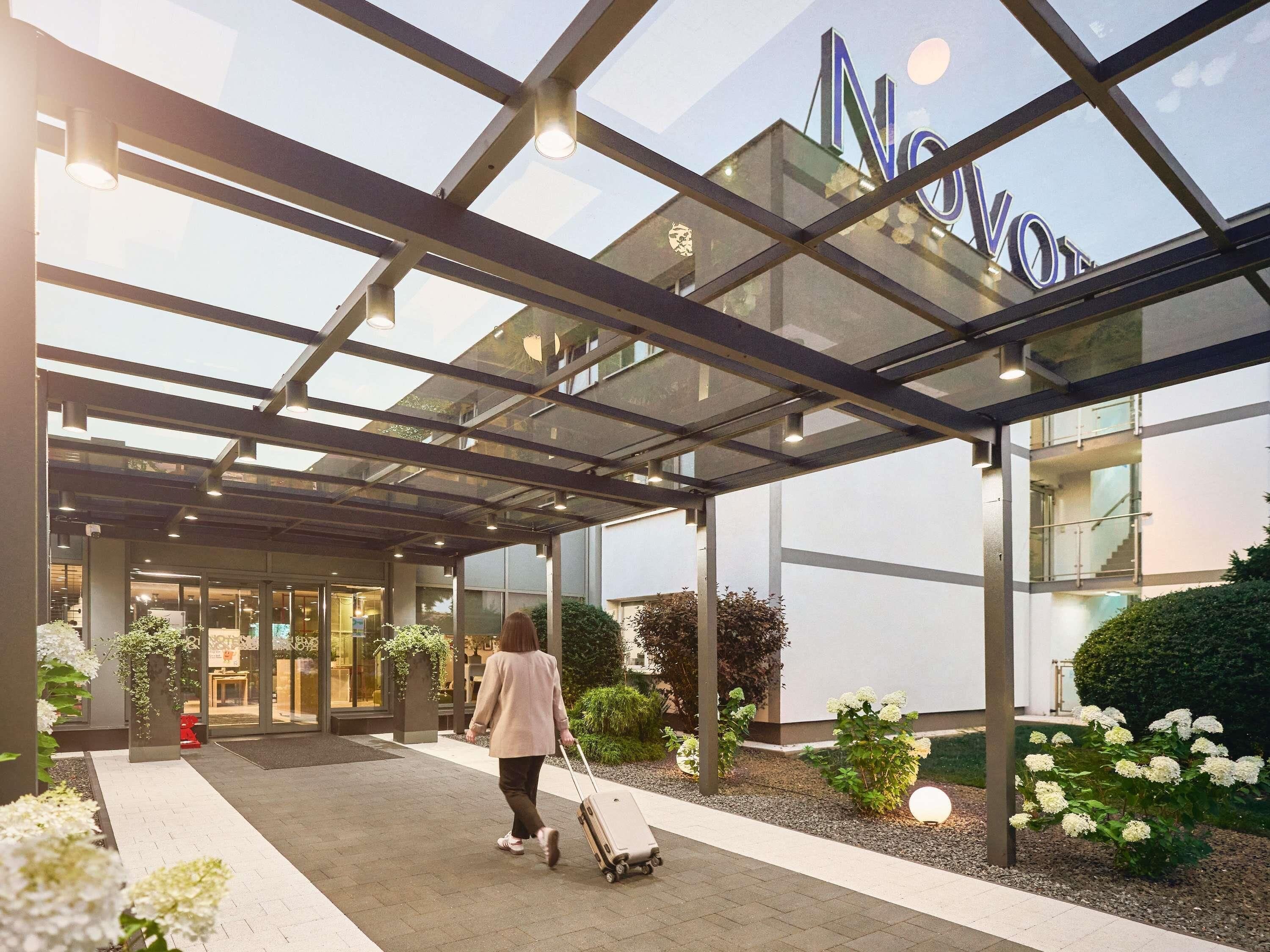 Entrada de hotel con toldo de cristal, una persona arrastrando una maleta y un jardín bien cuidado con flores blancas.