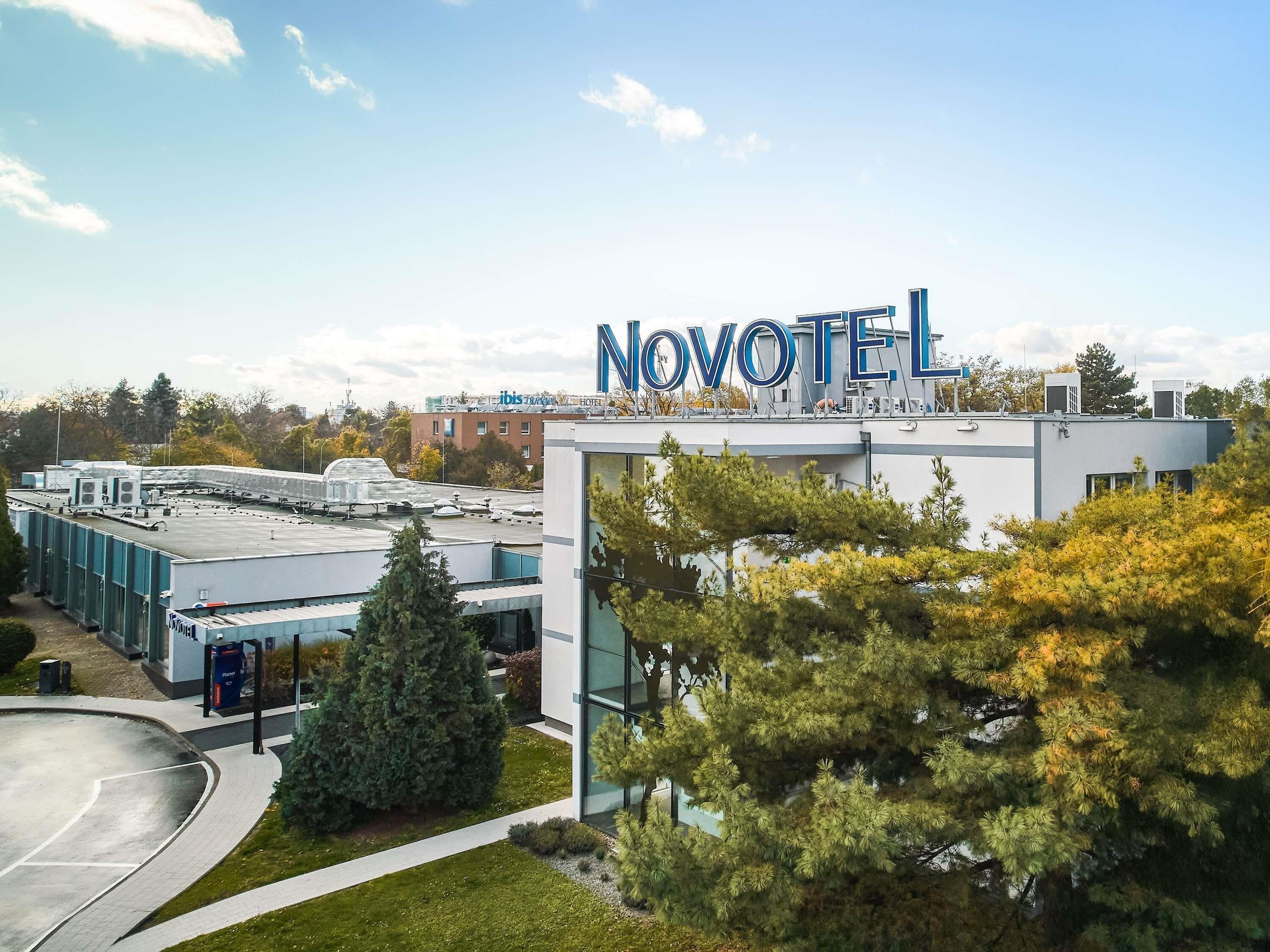 The Novotel hotel with a modern design, featuring a prominent 'Novotel' sign on the roof, surrounded by greenery and a clear sky.
