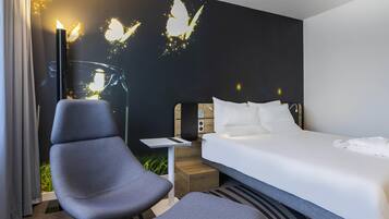 A hotel room with a bed, two chairs, a nightstand, a lamp, and a wall with butterfly projections.