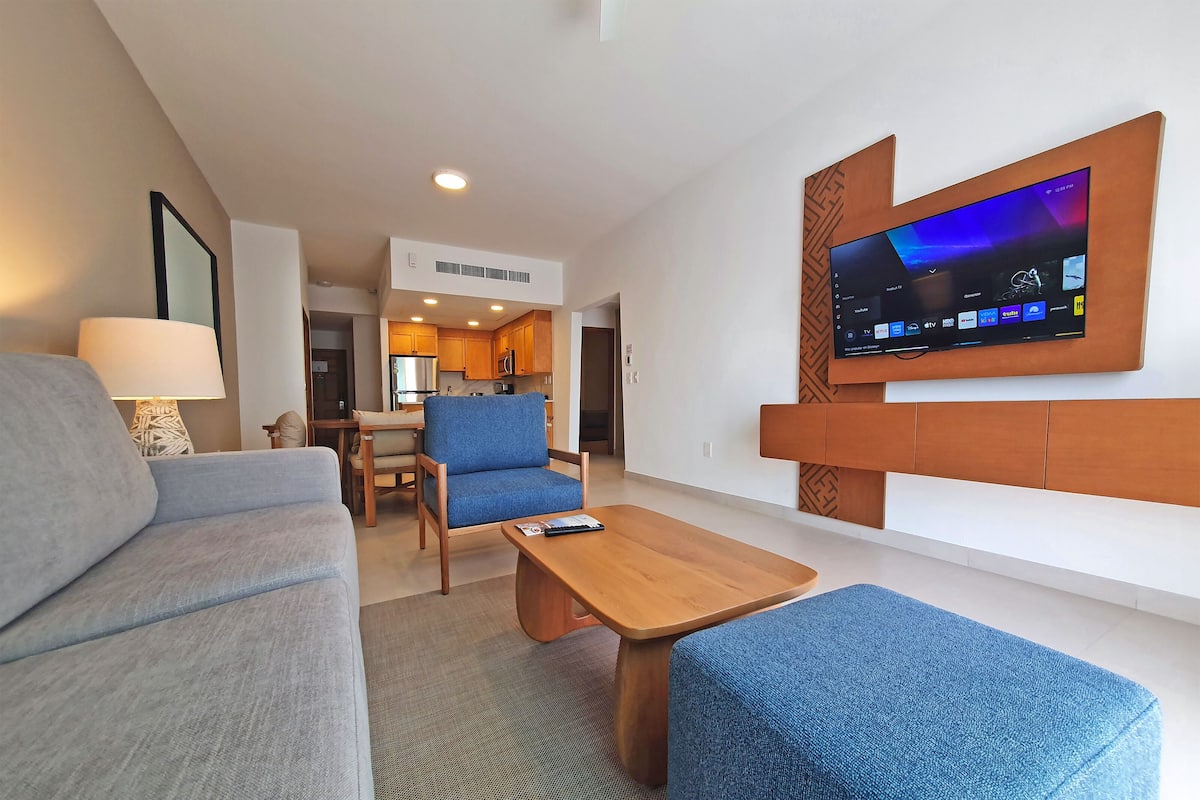 Een moderne hotelkamer met een wandgemonteerde flatscreen televisie, een bank, een blauwe fauteuil, een houten salontafel en een eethoek op de achtergrond.
