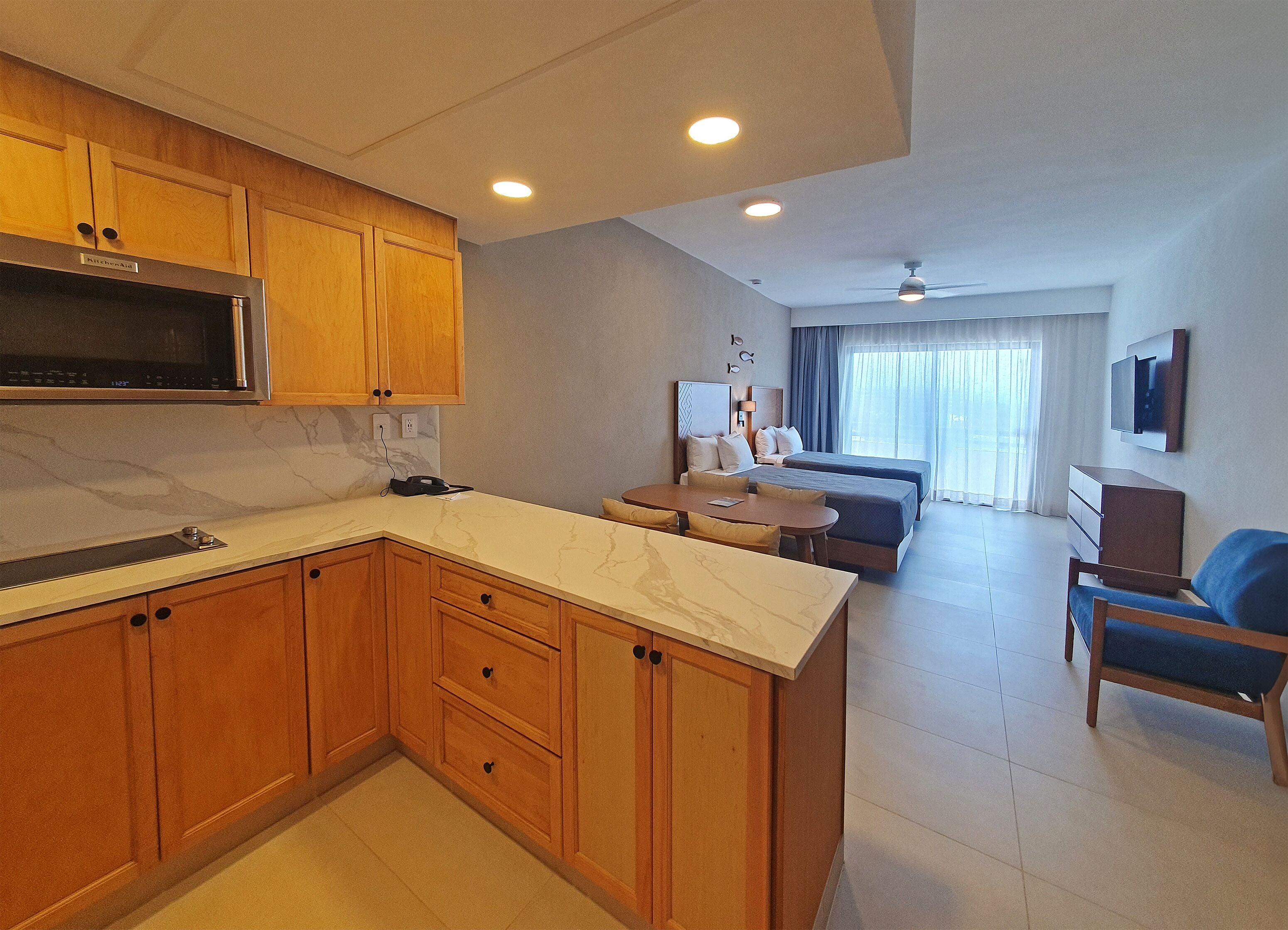 A modern kitchen with wooden cabinets and a marble countertop, a dining area with a table and chairs, a sofa, and a TV mounted on the wall.