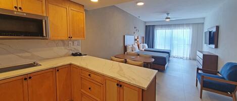A modern kitchen with wooden cabinets and a marble countertop, a dining area with a table and chairs, a sofa, and a TV mounted on the wall.