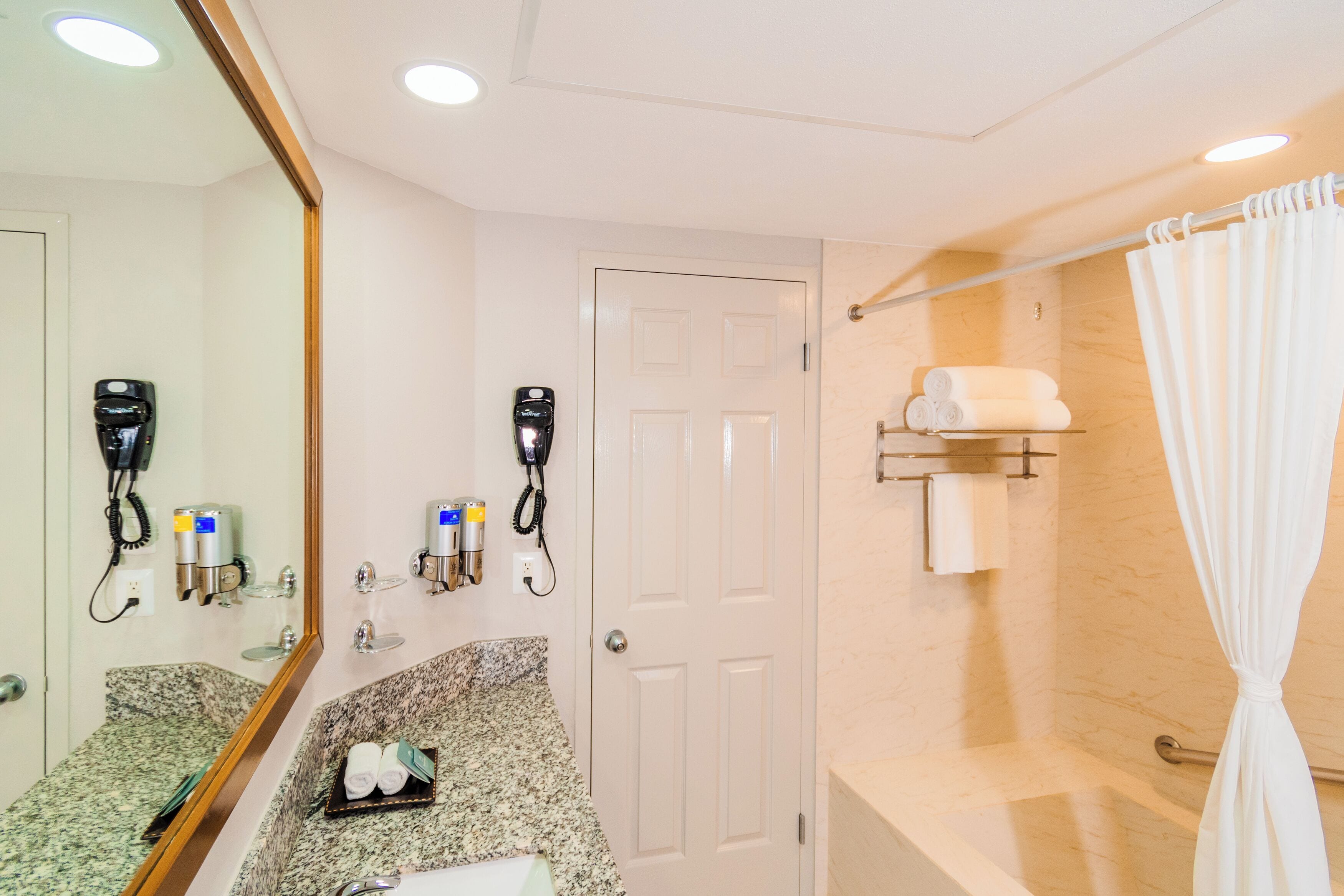 Une salle de bain dotée d’un comptoir en granit, d’un miroir, d’un sèche-cheveux, d’un porte-serviettes et d’un rideau de douche.