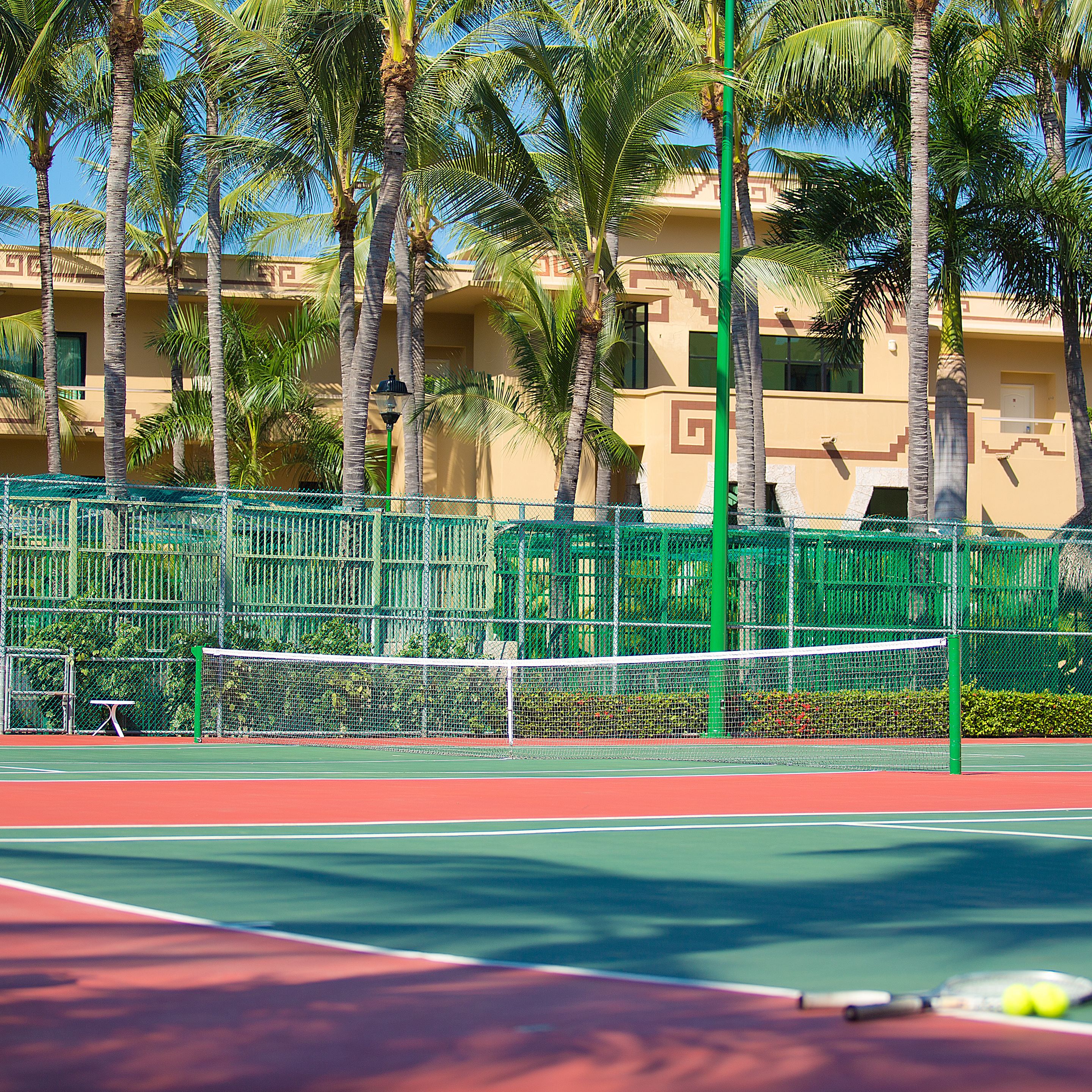 A tennis court with a net, surrounded by a green fence and palm trees.