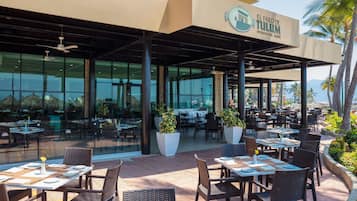 An outdoor dining area with tables and chairs, a large potted plant, and a building with a sign reading "El Faro de Tulum International Cuisine".