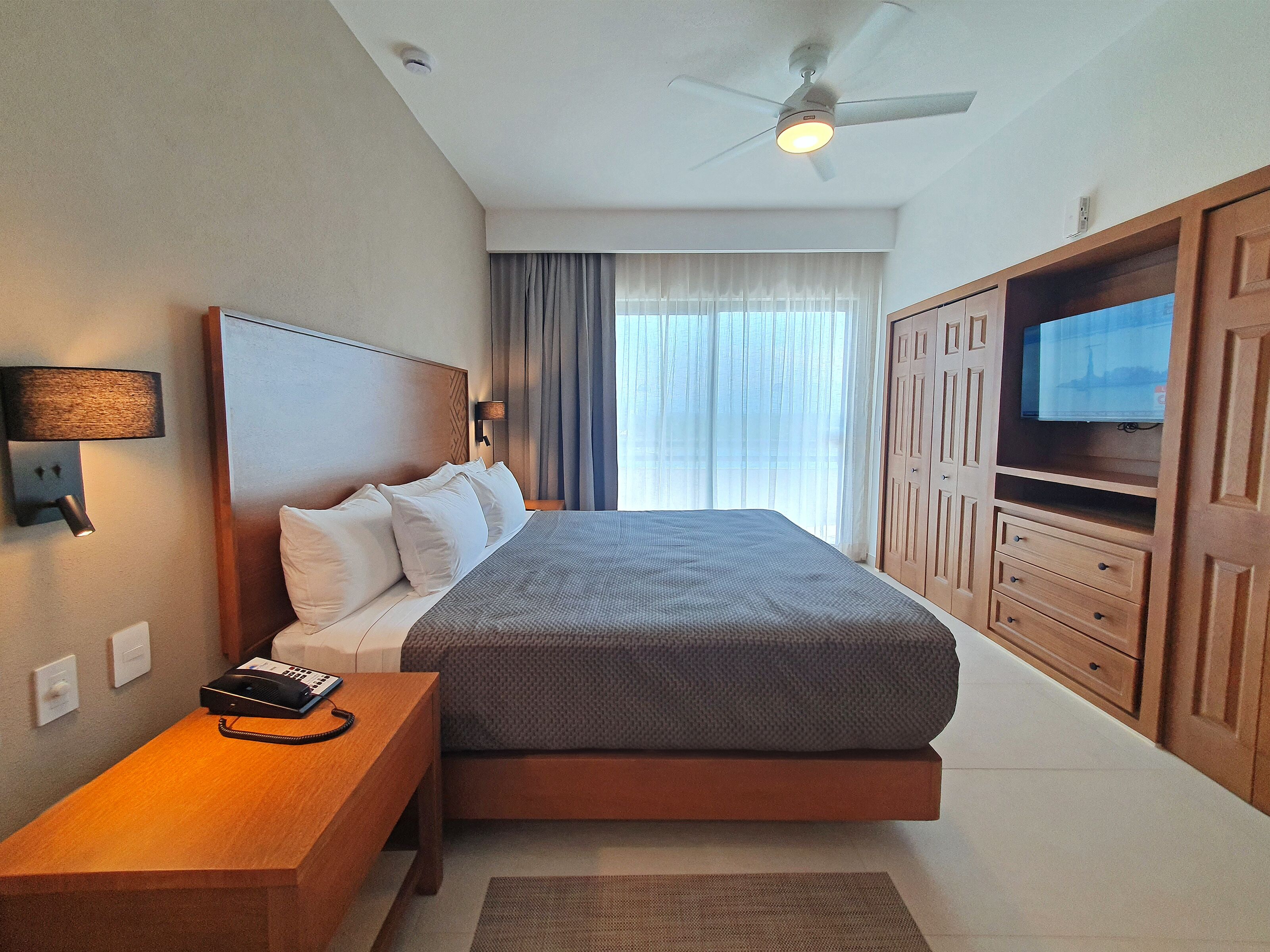 A hotel room with a large bed, a wooden nightstand, a ceiling fan, a TV, and a window with curtains.